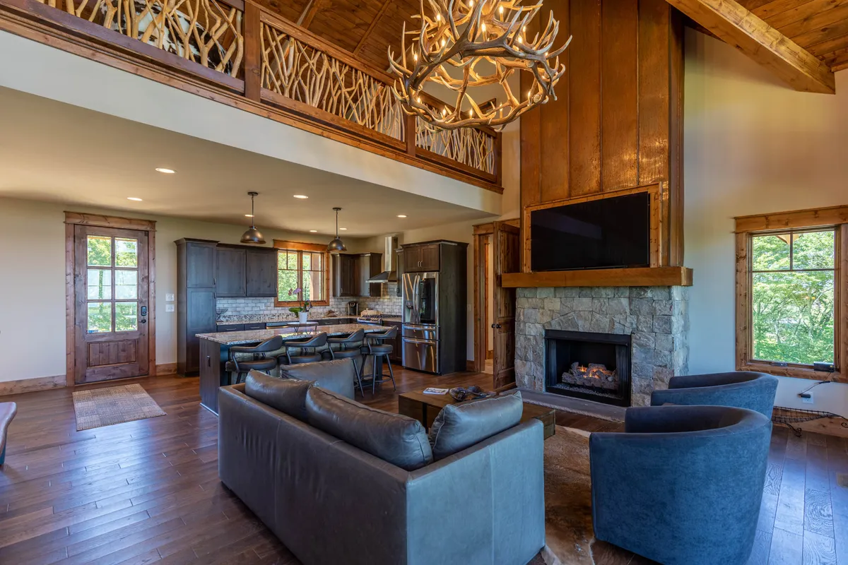 A warm and inviting open-concept space featuring a striking fireplace, antler chandelier, central kitchen island, and a spacious dining table. The living, dining, and kitchen areas flow naturally together, with comfortable seating arranged around the fireplace and ample natural light highlighting the modern yet homey finishes.