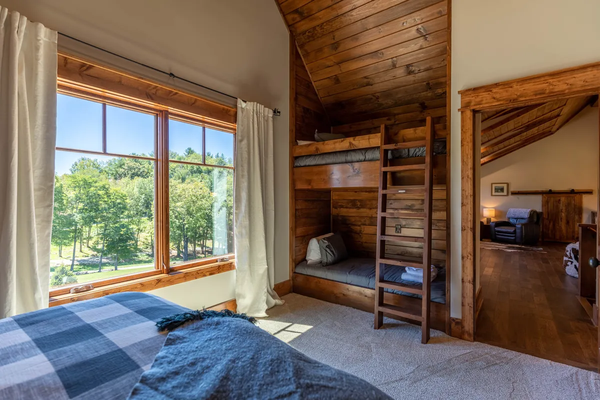 A comfortable bedroom with large windows framing green views, plaid bedding on bunk beds, and inviting natural light. The space feels playful yet restful, ideal for families or children.