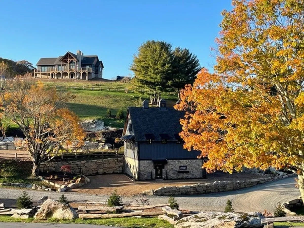 A welcoming exterior view featuring a large wooden house and a stone building set against vibrant autumn foliage. The foreground suggests space for gatherings, while the backdrop hints at nearby trails and outdoor adventure.