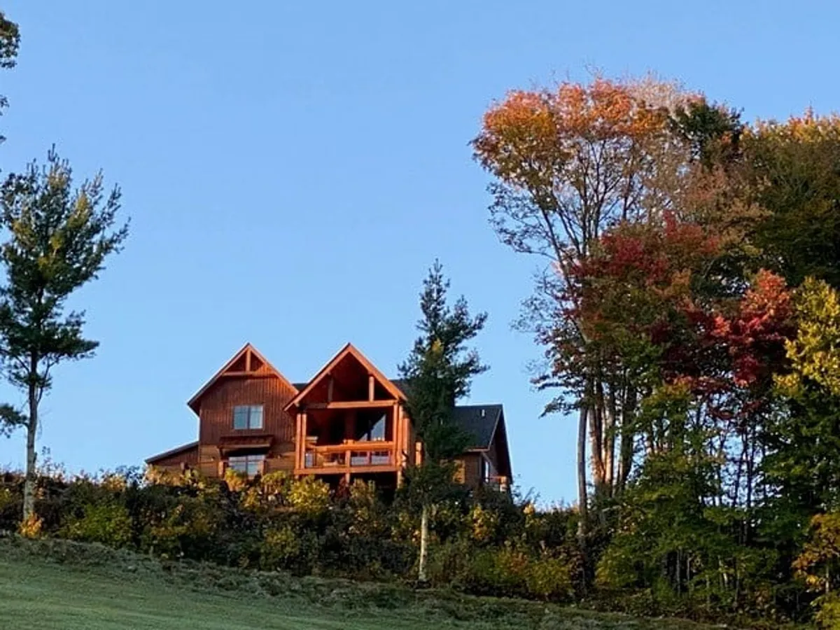 The wooden house is surrounded by tall trees and set against a backdrop of a clear blue sky. The scene evokes tranquility and hints at the property’s proximity to hiking trails and natural surroundings ideal for exploration and adventure.