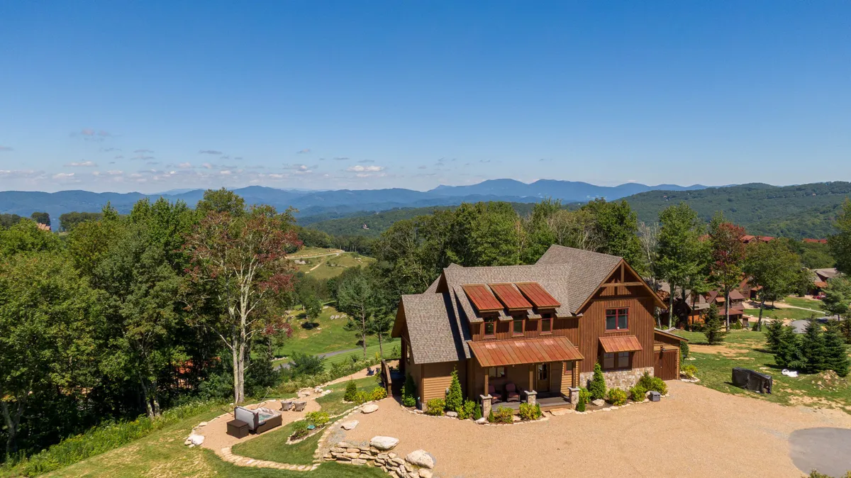 The image showcases a charming rustic cabin perched atop a lush, landscaped knoll, surrounded by sweeping mountain vistas. The house features a spacious porch perfect for sunrise coffee or sunset cocktails, with plenty of space in the landscaped yard for gatherings, relaxation, or stargazing under clear North Carolina skies.