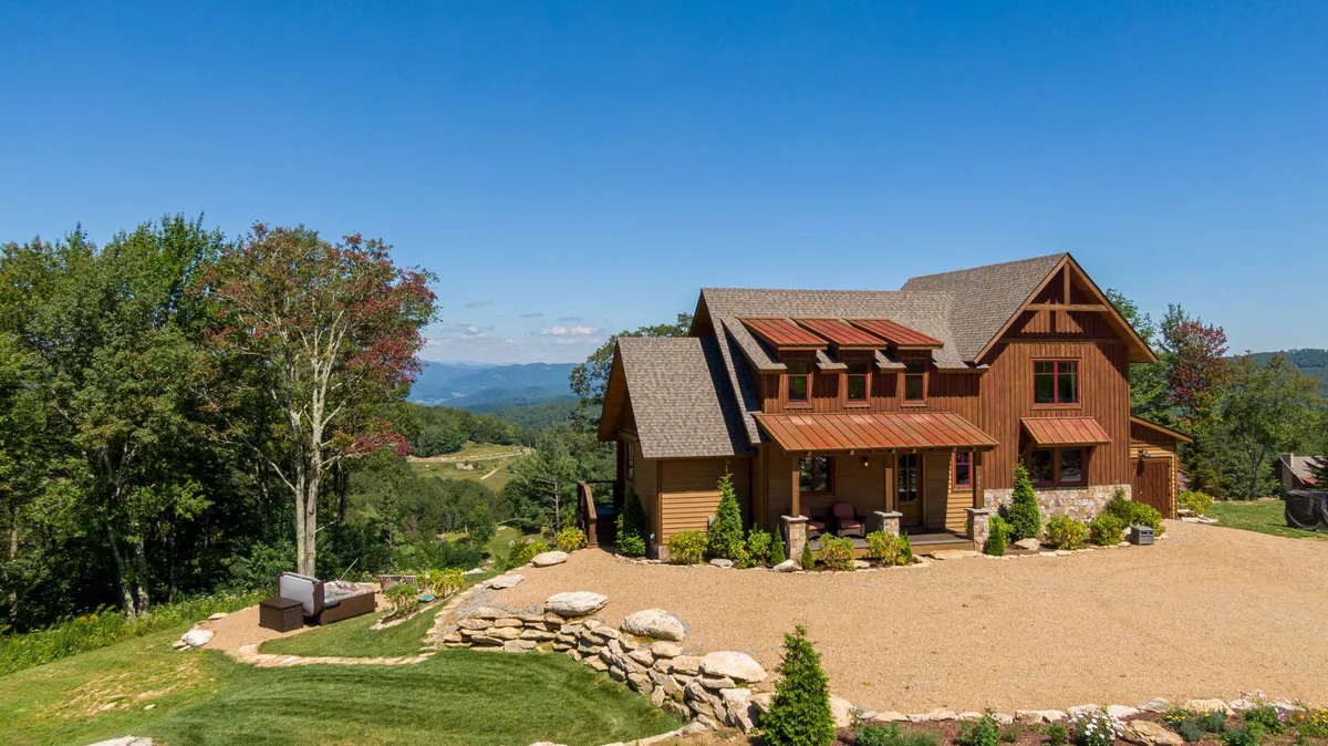 The image displays the home's wooden facade nestled amid a landscaped yard, with expansive mountain views stretching into the background. The atmosphere is serene and welcoming, suggesting an inviting retreat surrounded by natural beauty.