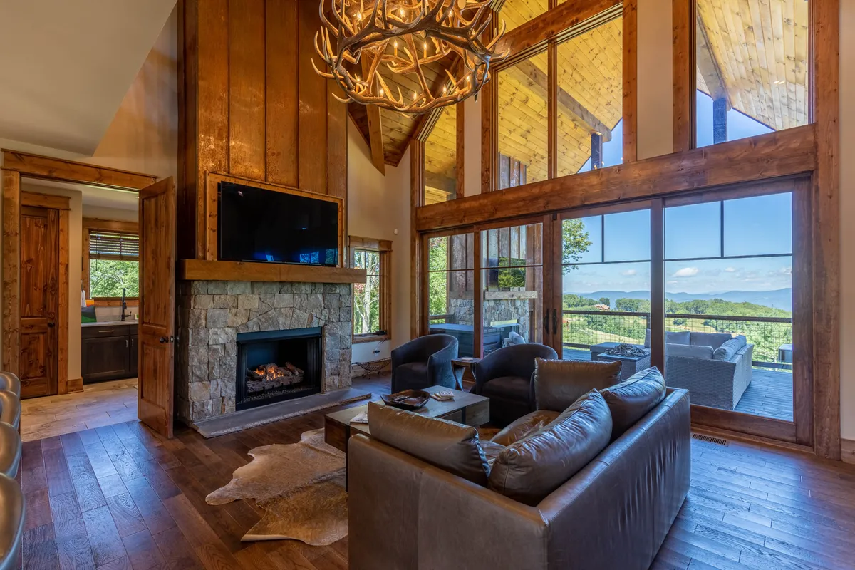 A cozy living room centered around a beautiful stone fireplace, with large windows offering scenic views and a statement chandelier overhead. Comfortable seating invites guests to relax by the fire or enjoy the mountain breeze from the adjacent deck.