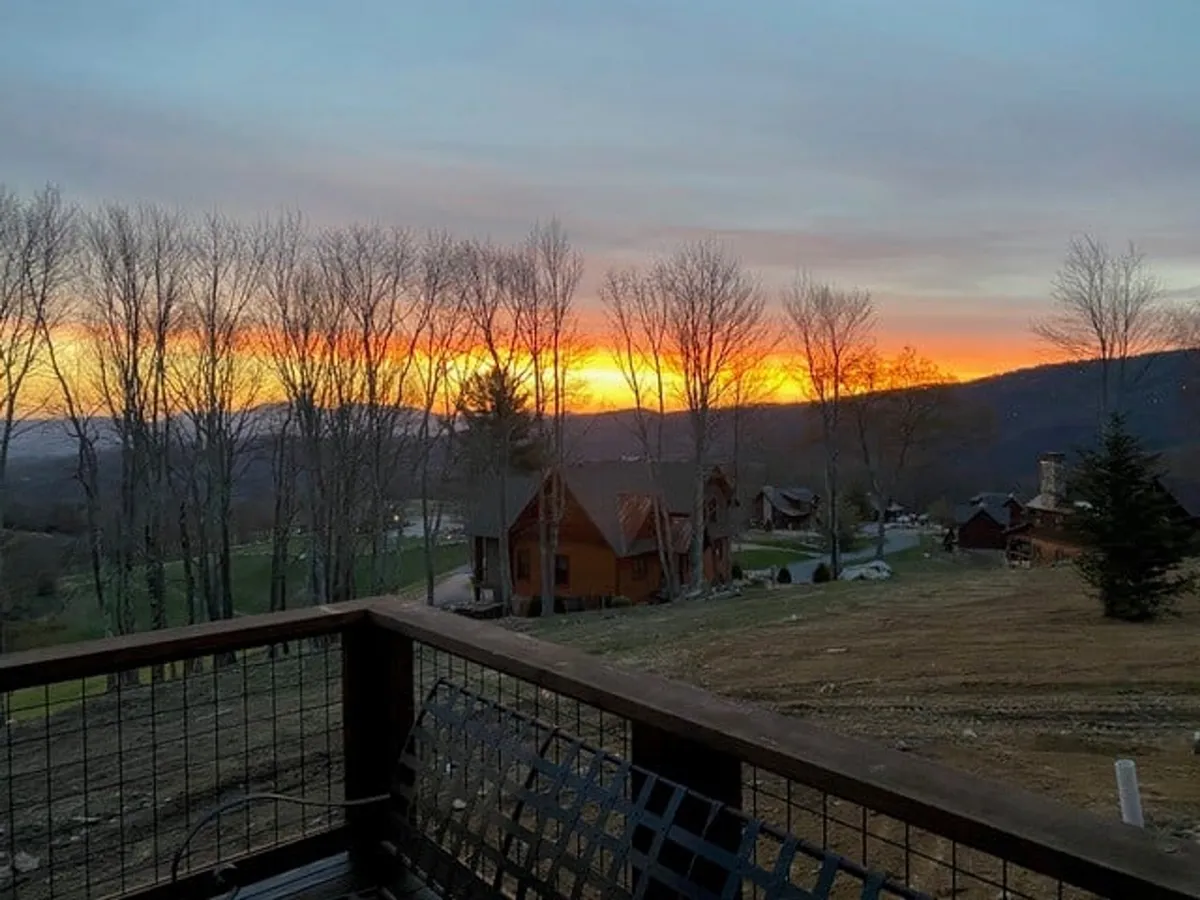 This scenic view image captures a stunning sunset over distant mountains and a tree-lined horizon, with cozy cabins nestled below. The atmosphere is peaceful and prime for evening activities like stargazing, taking in the view, or capturing photos with friends.