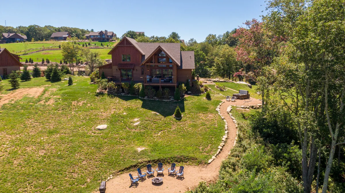 A spacious wooden house is nestled among lush greenery, with a prominent fire pit area and comfortable seating arranged on a stone patio. A pathway leads from the house to the cozy outdoor gathering spot, inviting families to enjoy evenings together surrounded by nature.