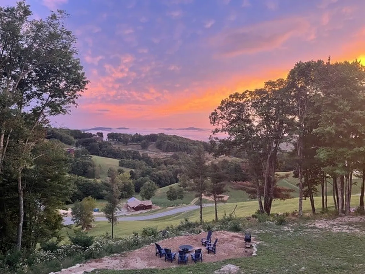 A seating area encircles a glowing fire pit, set against the backdrop of rolling hills, a colorful sunset, and distant trees. The tranquil atmosphere invites groups to gather, relax, and share stories as the day ends.
