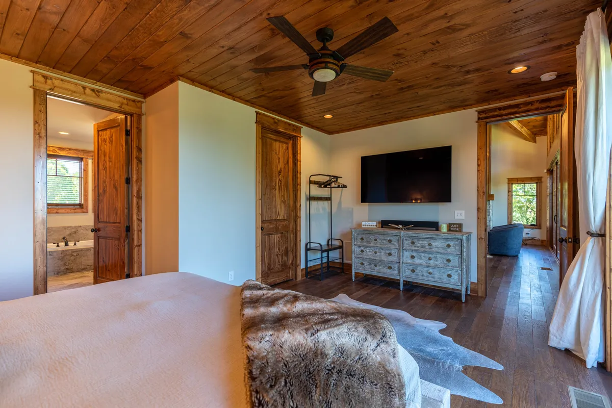A cozy bedroom featuring a bed with a plush throw blanket, a wooden ceiling, a TV unit, and double doors leading to a private bathroom. Soft natural light fills the room, creating a peaceful retreat.
