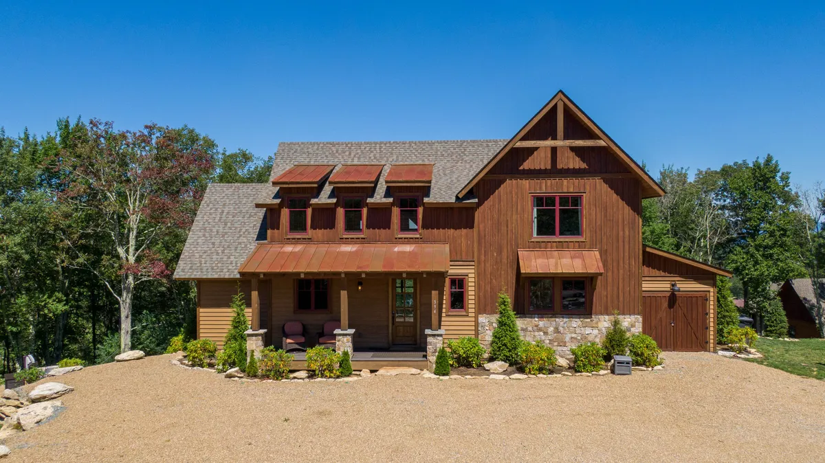 This exterior shot features the home's wooden facade with stone accents, a cozy front porch seating area, and a lush, landscaped garden. The scene suggests moments of relaxation, whether enjoying a morning coffee or toasting the sunset on the porch amidst nature.