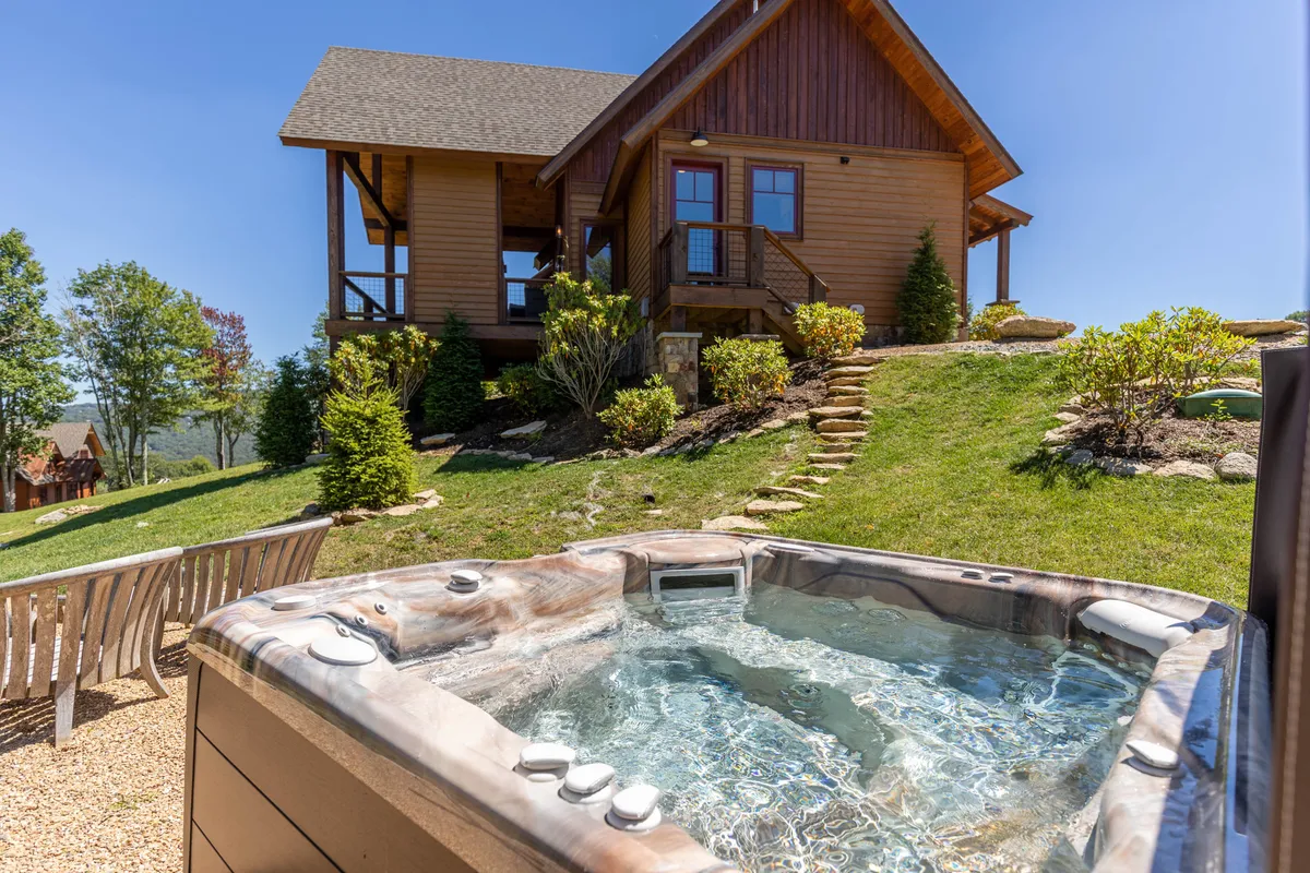 A tranquil exterior scene with a hot tub beside a wooden house, landscaped gardens, and ample space for outdoor socializing. Guests are invited to relax in the hot tub, enjoy nature walks, or gather for barbecues and stargazing.
