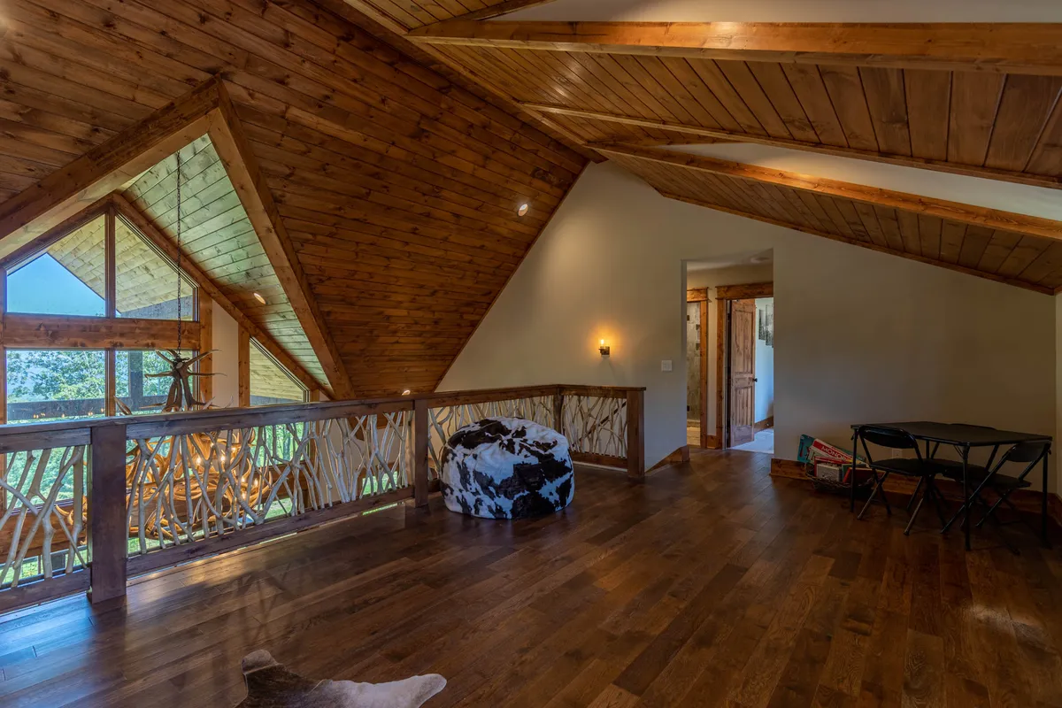 A loft space with exposed wooden beams, large windows overlooking natural scenery, a cozy seating area with bean bags, and decorative railing. The space is bathed in natural light and feels open yet inviting.