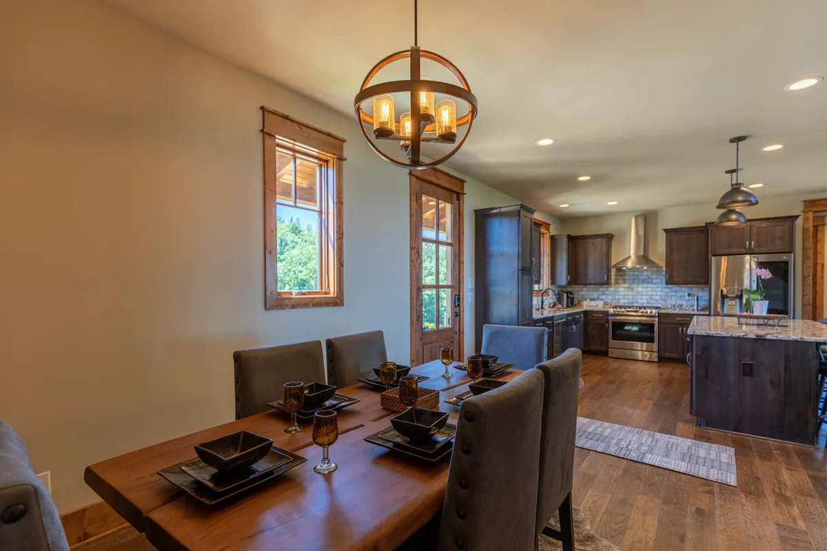 A welcoming dining area set for a gathering, with a chandelier overhead, a view into the kitchen, and windows filtering in natural light. The layout encourages togetherness, whether for a family dinner or a casual breakfast with friends.