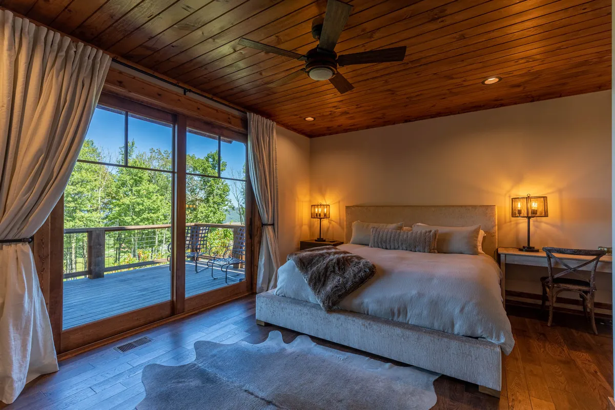 A spacious primary bedroom with a large bed adorned with decorative pillows, floor-to-ceiling windows letting in abundant natural light, a wooden ceiling, and stylish lighting fixtures. The room opens to a private deck, inviting relaxation with nature views.